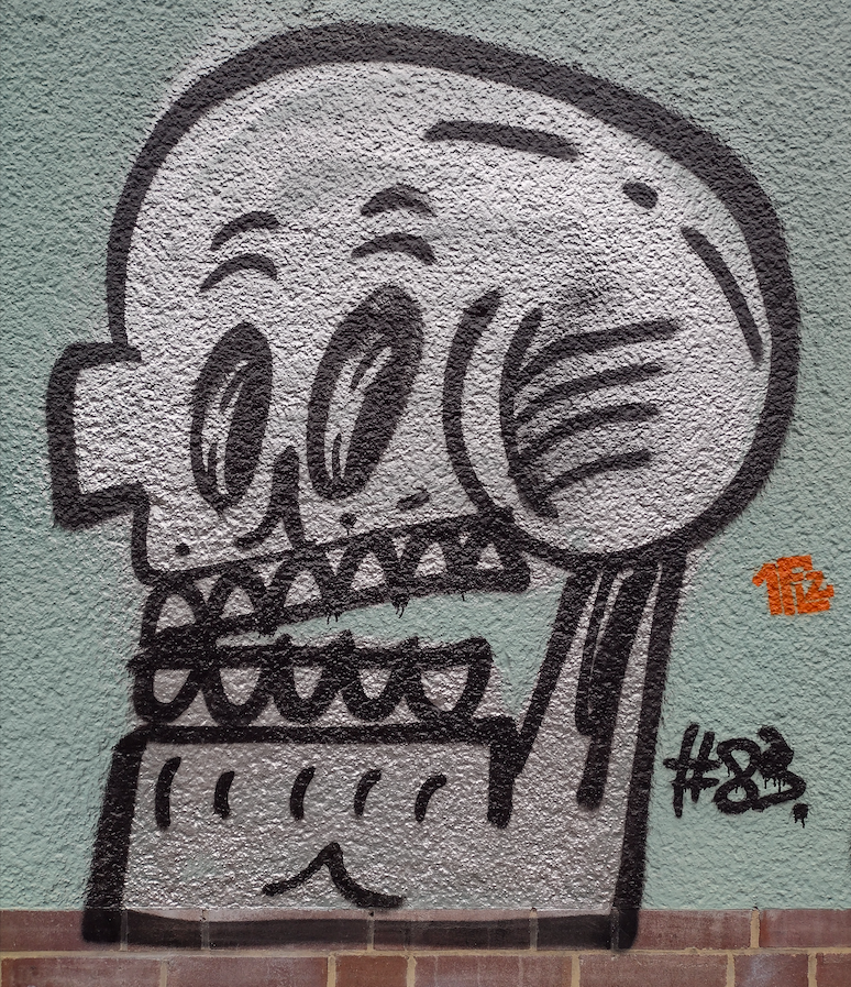Skull number 83. It is a prime example (pun intended) of this style of graffiti, thickly outlined in black and filled with silver spray paint. It's a two-dimensional, almost Picassoesque caricature of a leftward facing skull, with a completely angular left ear and jaw, in stark contrast to the roundness of the top of the skull and the right ear. There is no depth to the mouth with its two flat rows of bared teeth (six above, five below), yet the skull appears surprisingly expressive. The artists seem to achieve this by adding (live) human facial features such as black irises within the elliptical eye holes that look cheekily sideways at the observer, and semi-circular "eyebrows" that give it an air of surprise. There are other squiggles and dots on the skull that add dimension.
