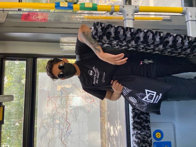 Vagrant dressed in all black, a black mask and black round sunglasses. A carabiner with keys hangs from a belt loop and a tote bag from the Deutsche Kinemathek (German film museum) hangs from their shoulder as they lean against a backrest in a tram, showing a tattooed forearm. Their t-shirt says "Saarbrigge, du Geiler!" because it is from Saarbrücken.
