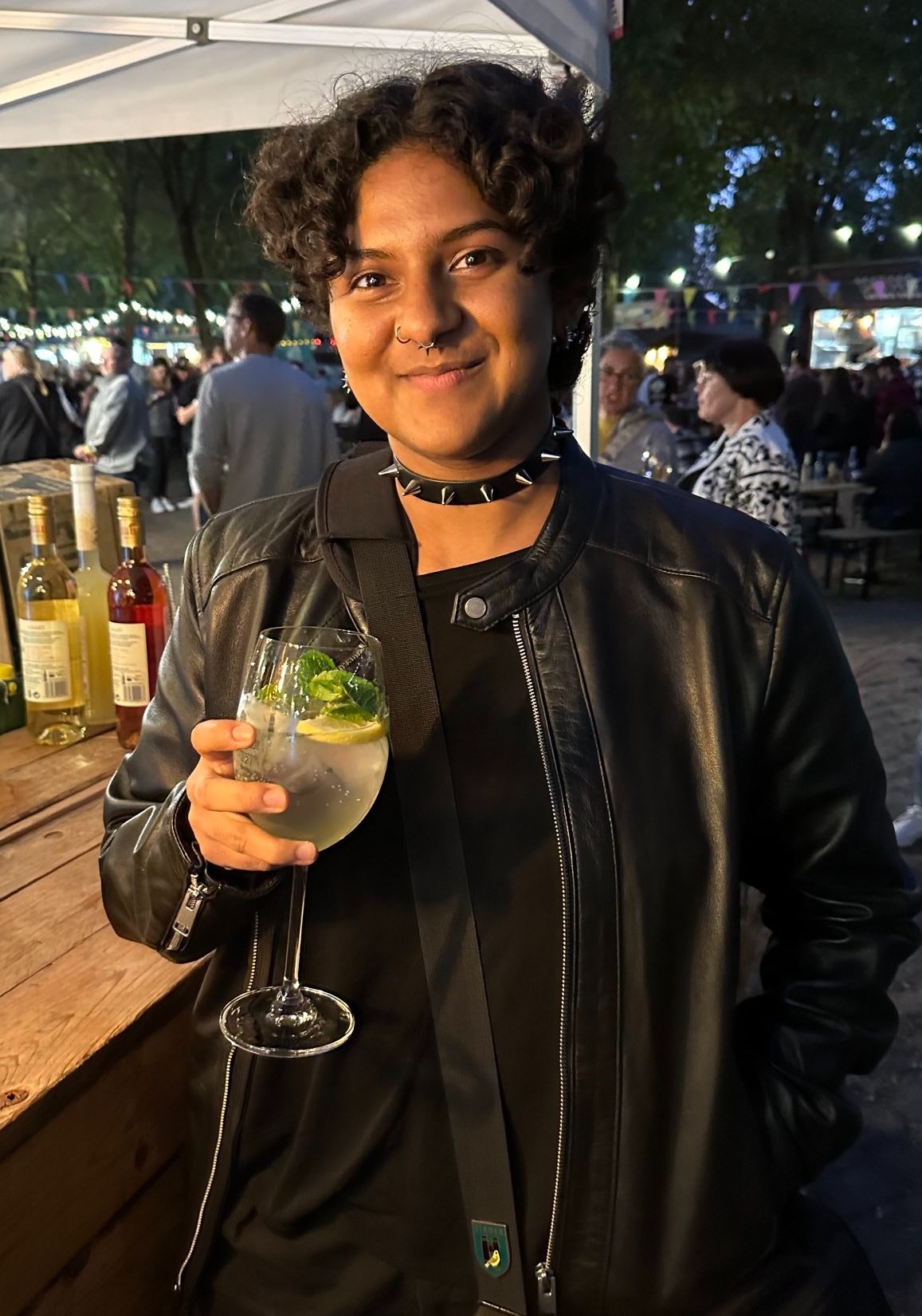 Vagrant, a brown-skinned person with short curly black hair, smiles at the camera. They are wearing all-black clothes, a black leather jacket, and a leather choker with spikes. Their hand holds a mocktail in a wine glass.